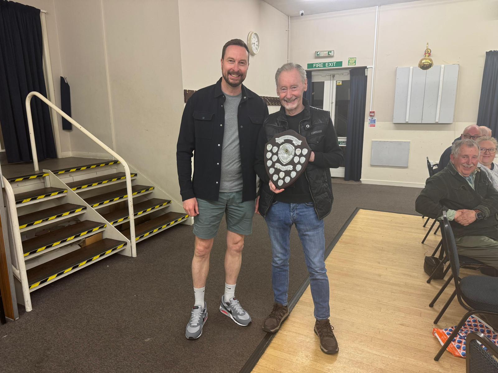 Two smiling older men standing in a small meeting room, one shaking hands with the other while the other holds a circular wooden shield trophy with small metal plates; they stand in front of a curtained window. The mood is celebratory, marking an award presentation for club find of the year.